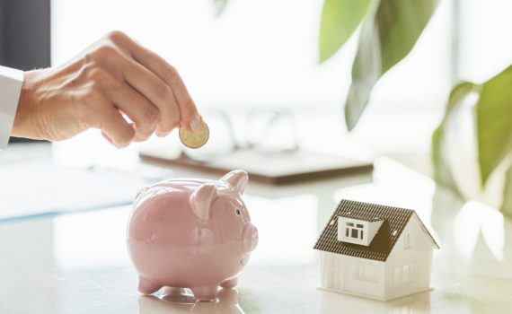 Hand putting money in White piggy bank with tiny house on white marble background and blurred bokeh with copy space. Concept for financial home loan or money saving for house buying, real estate and insurance